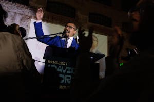 Photo of David Morales in a blue suit raising his hand in the air while speaking into a microphone. Silhouettes of outreached hands are in front of him.