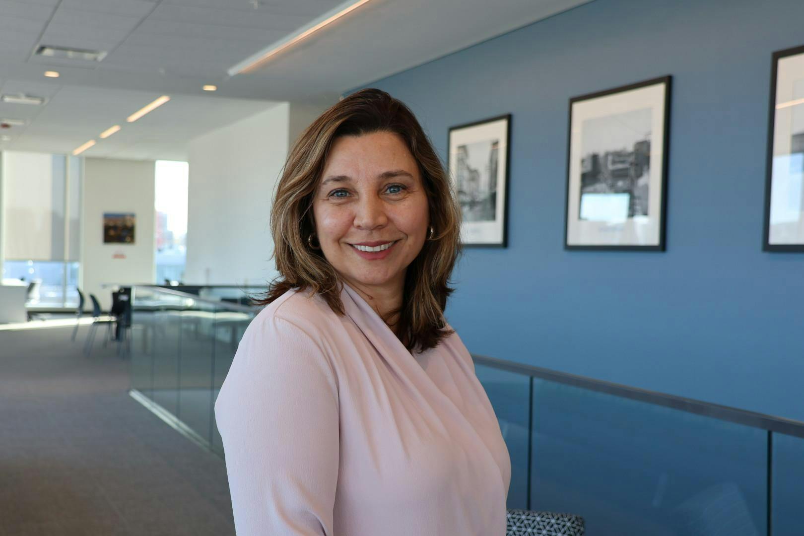 Sandra Smith smiles to the camera in a pink blouse. Behind her, framed photographs stretch along a blue wall.