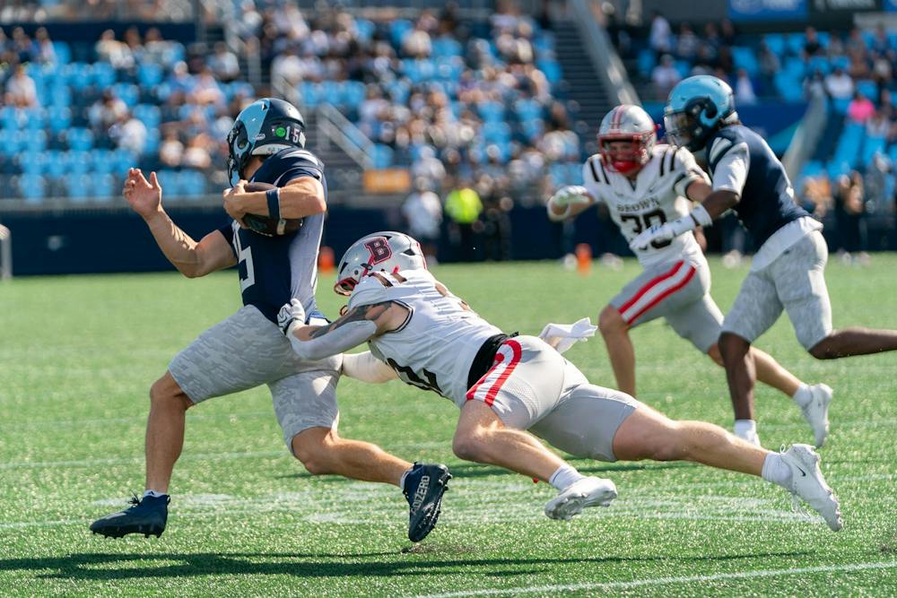 A photo of a Brown football player diving to tackle a URI player running with the ball. 

