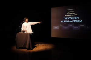 Terrence Nance in a white long sleeve shirt stands behind a podium with a laptop and a microphone in a dimly lit room, gesturing with his left arm to the large screen. The screen displays the text: "SUN RA says Black people need The sound of our own MYTHS," followed by the main title "THE CONCEPT ALBUM as CINEMA."