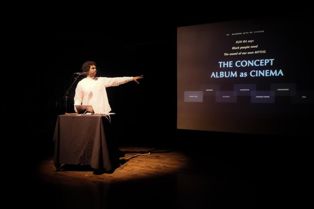Terrence Nance in a white long sleeve shirt stands behind a podium with a laptop and a microphone in a dimly lit room, gesturing with his left arm to the large screen. The screen displays the text: "SUN RA says Black people need The sound of our own MYTHS," followed by the main title "THE CONCEPT ALBUM as CINEMA."