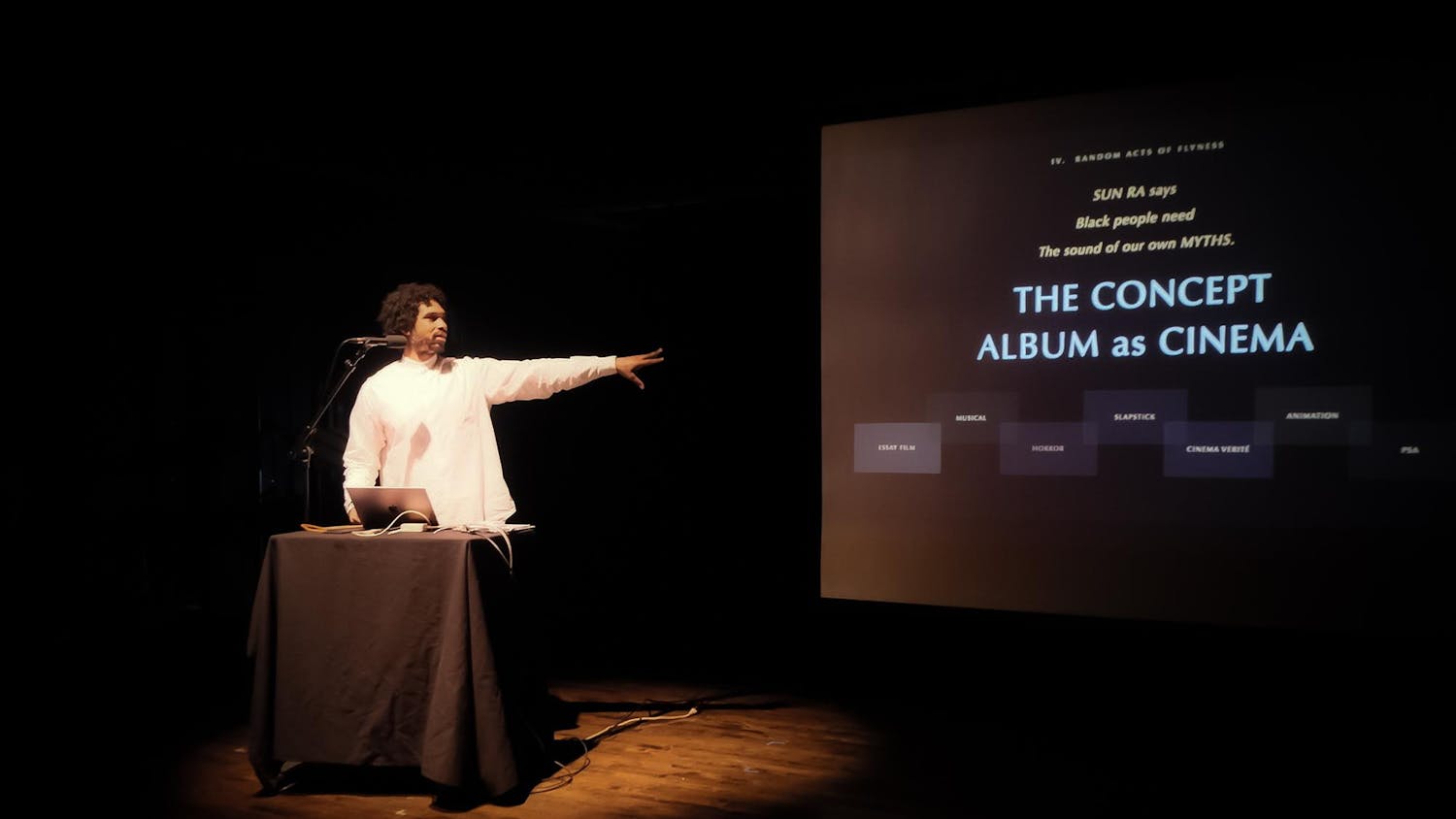 Terrence Nance in a white long sleeve shirt stands behind a podium with a laptop and a microphone in a dimly lit room, gesturing with his left arm to the large screen. The screen displays the text: "SUN RA says Black people need The sound of our own MYTHS," followed by the main title "THE CONCEPT ALBUM as CINEMA."