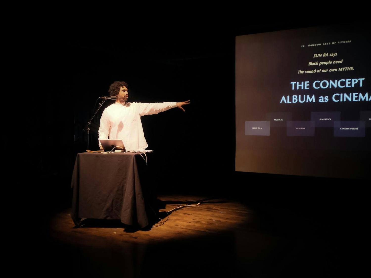 Terrence Nance in a white long sleeve shirt stands behind a podium with a laptop and a microphone in a dimly lit room, gesturing with his left arm to the large screen. The screen displays the text: "SUN RA says Black people need The sound of our own MYTHS," followed by the main title "THE CONCEPT ALBUM as CINEMA."