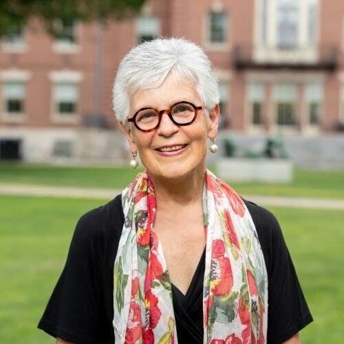 Janet Cooper Nelson smiles in a headshot. She has short silver hair, and is wearing a floral scarf with tortoiseshell glasses.