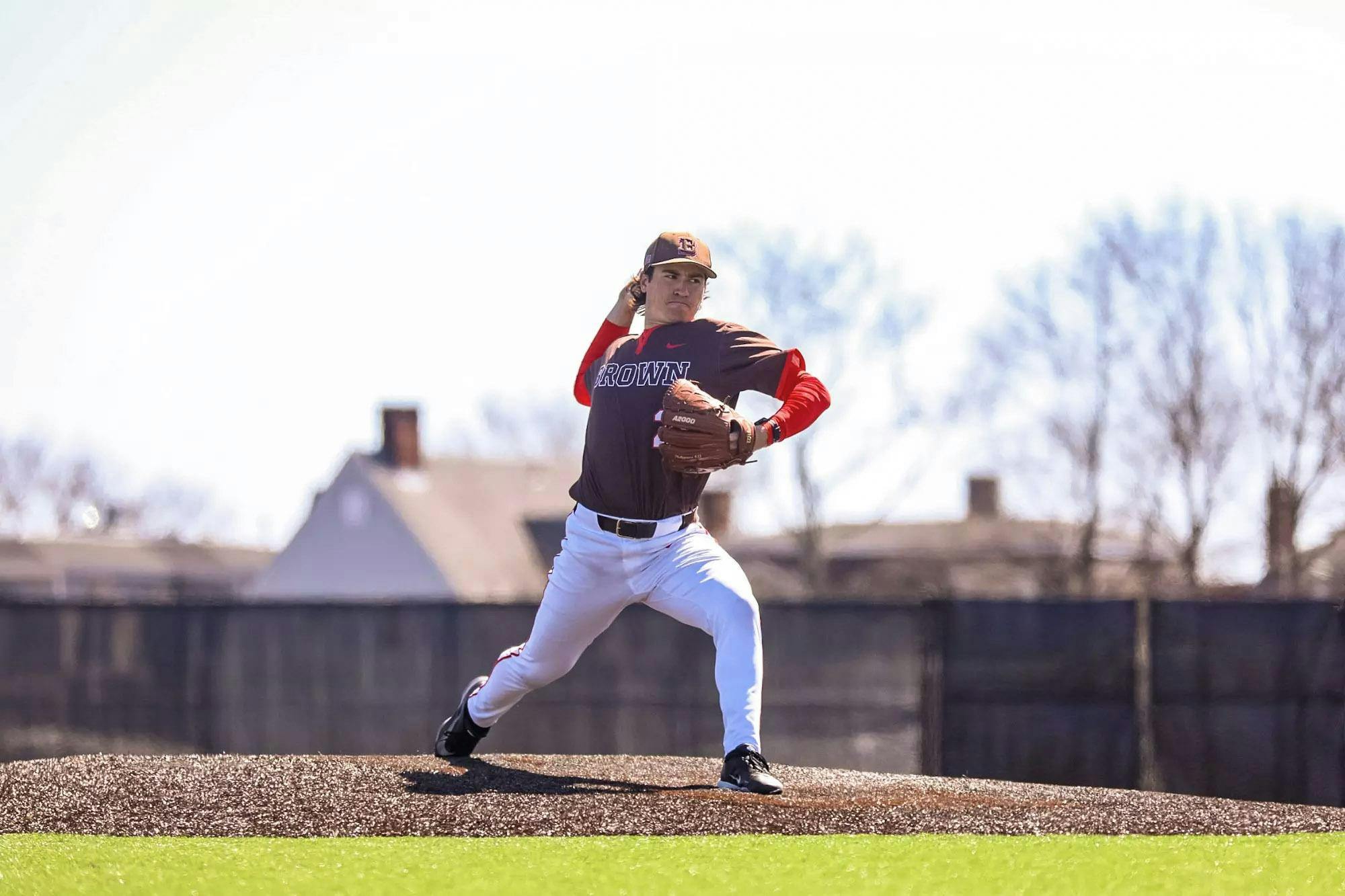 A photo of a baseball player wearing a Brown University jersey pitching a ball on the pitching mound during a sunny day. 