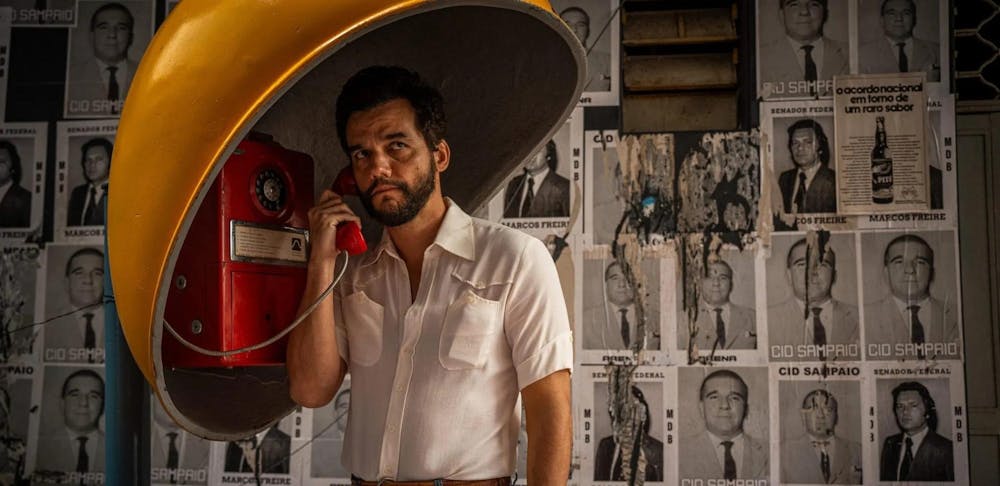 Still from the film of a concerned Alves in a white short sleeved button up with dark brown hair and a beard calling on a red payphone. 