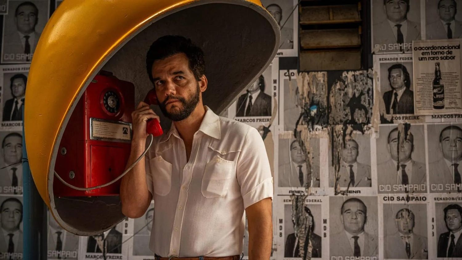 Still from the film of a concerned Alves in a white short sleeved button up with dark brown hair and a beard calling on a red payphone.