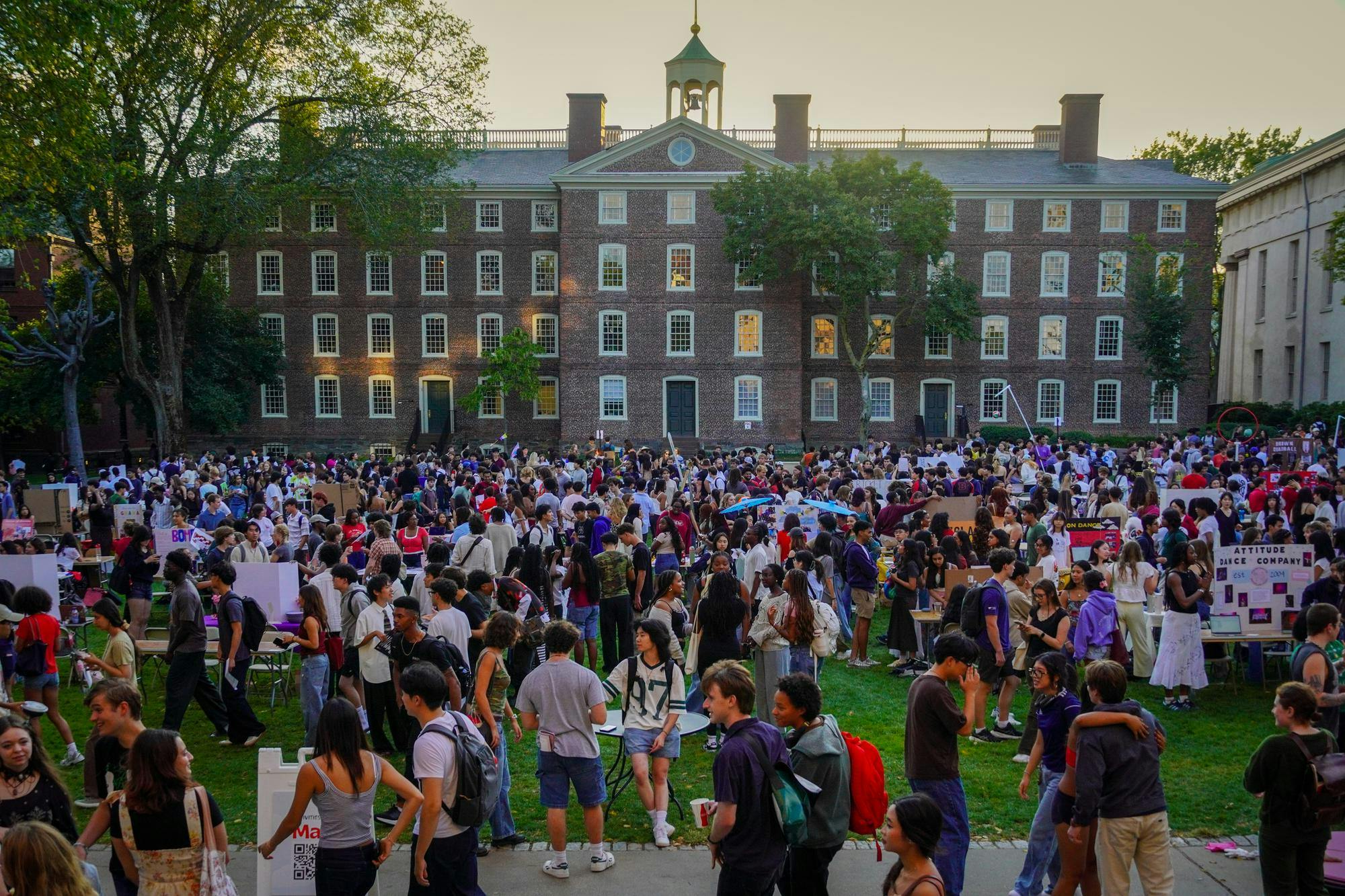 A large crowd of students stands on a large patch of grass in front of a University building. Various poster boards can be spotted within the crowd.