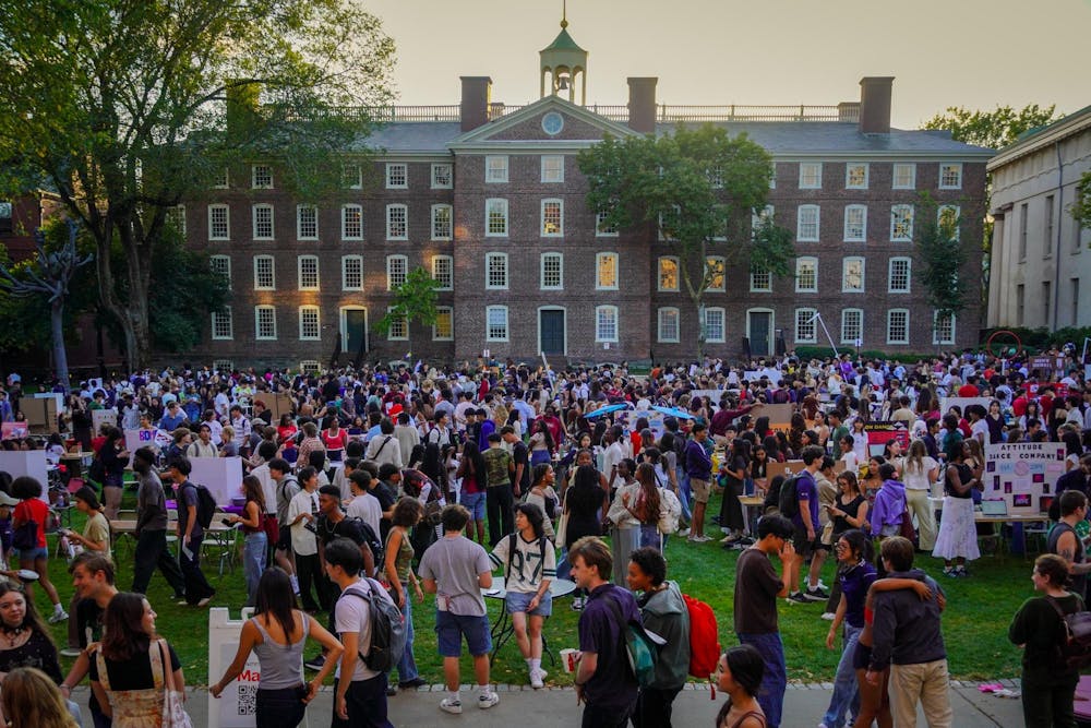A large crowd of students stands on a large patch of grass in front of a University building. Various poster boards can be spotted within the crowd.