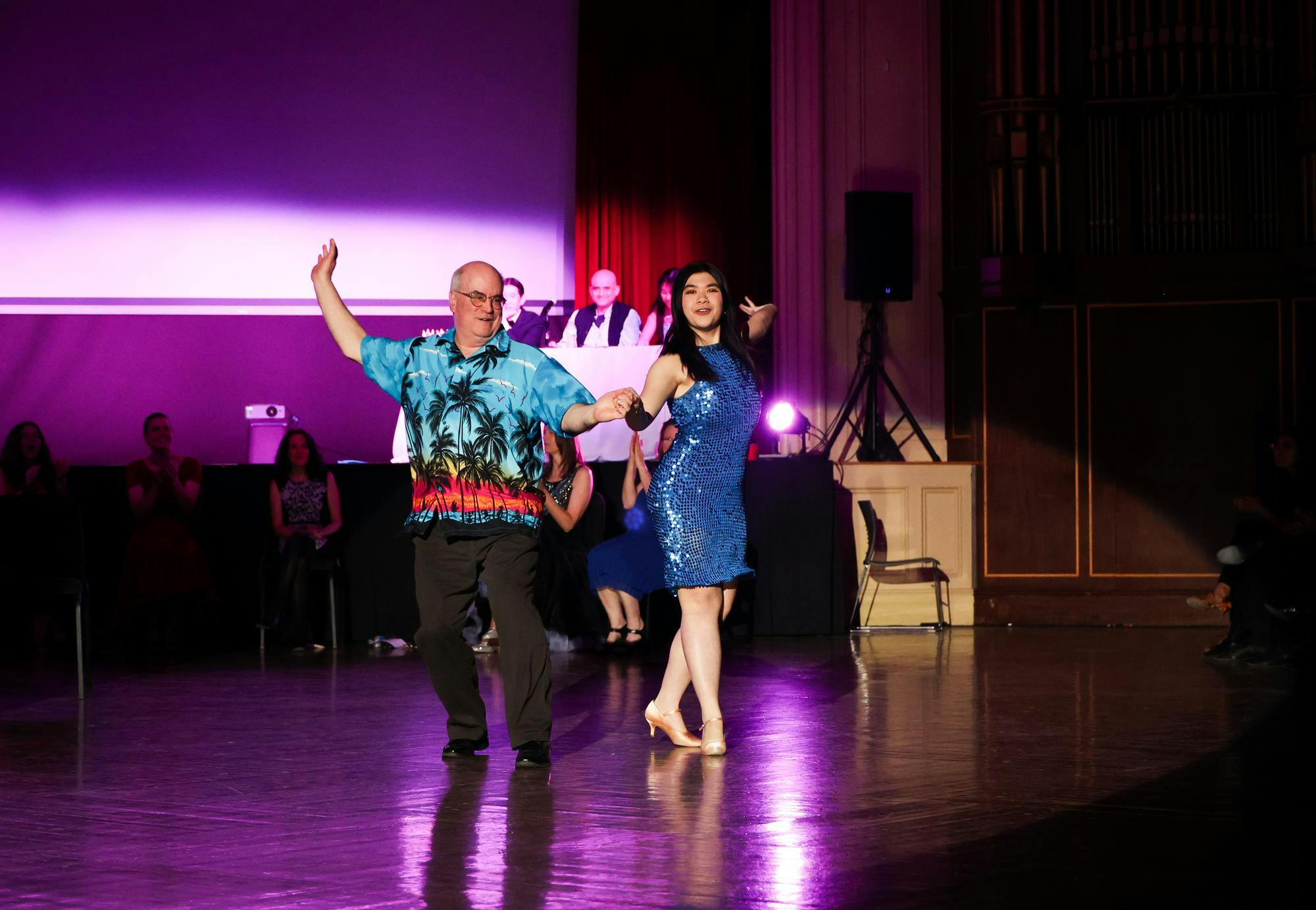A man in a Hawaiian shirt dances with a girl in a sparkly blue dress.