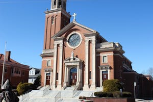 A photo of a red brick building with white stone details and dark brown wooden doors featuring a statue of a woman on top of the door. A large circular window is right above the door with crosses on the apex of the building.