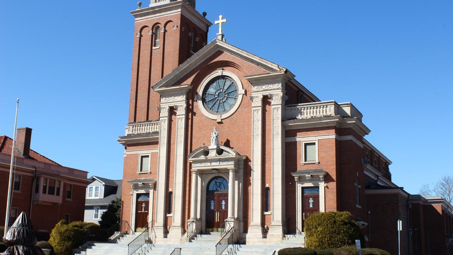 A photo of a red brick building with white stone details and dark brown wooden doors featuring a statue of a woman on top of the door. A large circular window is right above the door with crosses on the apex of the building.