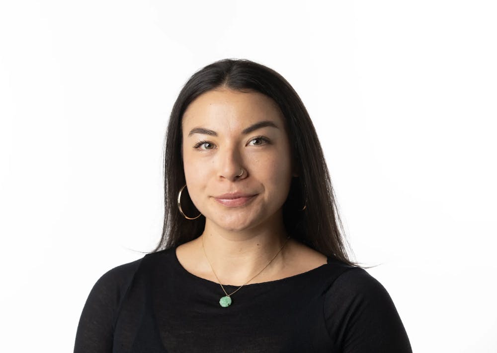 Profile photo of Anna Martin. A woman in her 30s with long straight dark brown hair, a black top, gold hoop earrings and a short necklace with a green pendant.