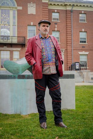 Photo of a man in a beret, a red jacket and multicolored shirt standing in front of a brick building.