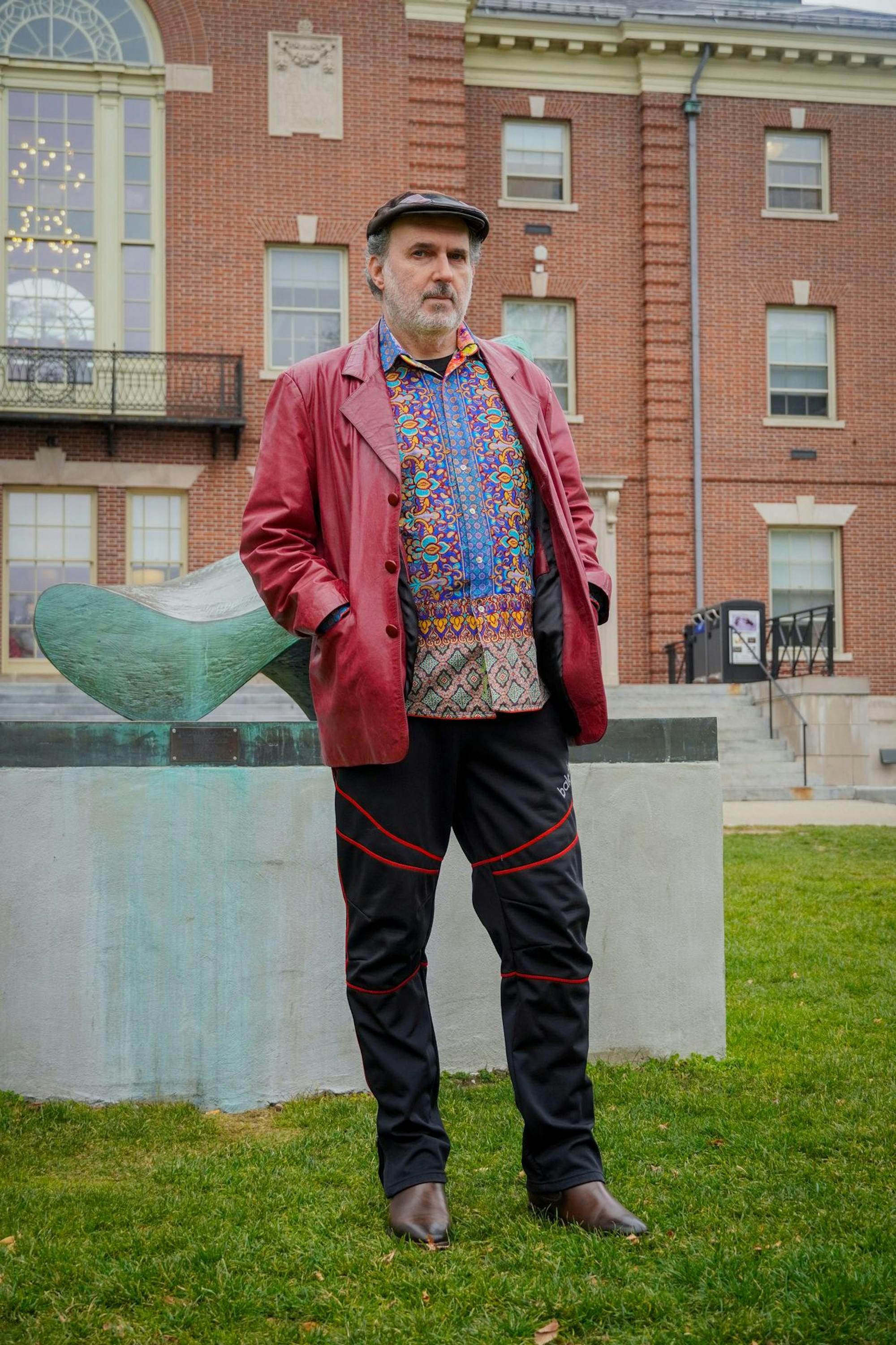 Photo of a man in a beret, a red jacket and multicolored shirt standing in front of a brick building.