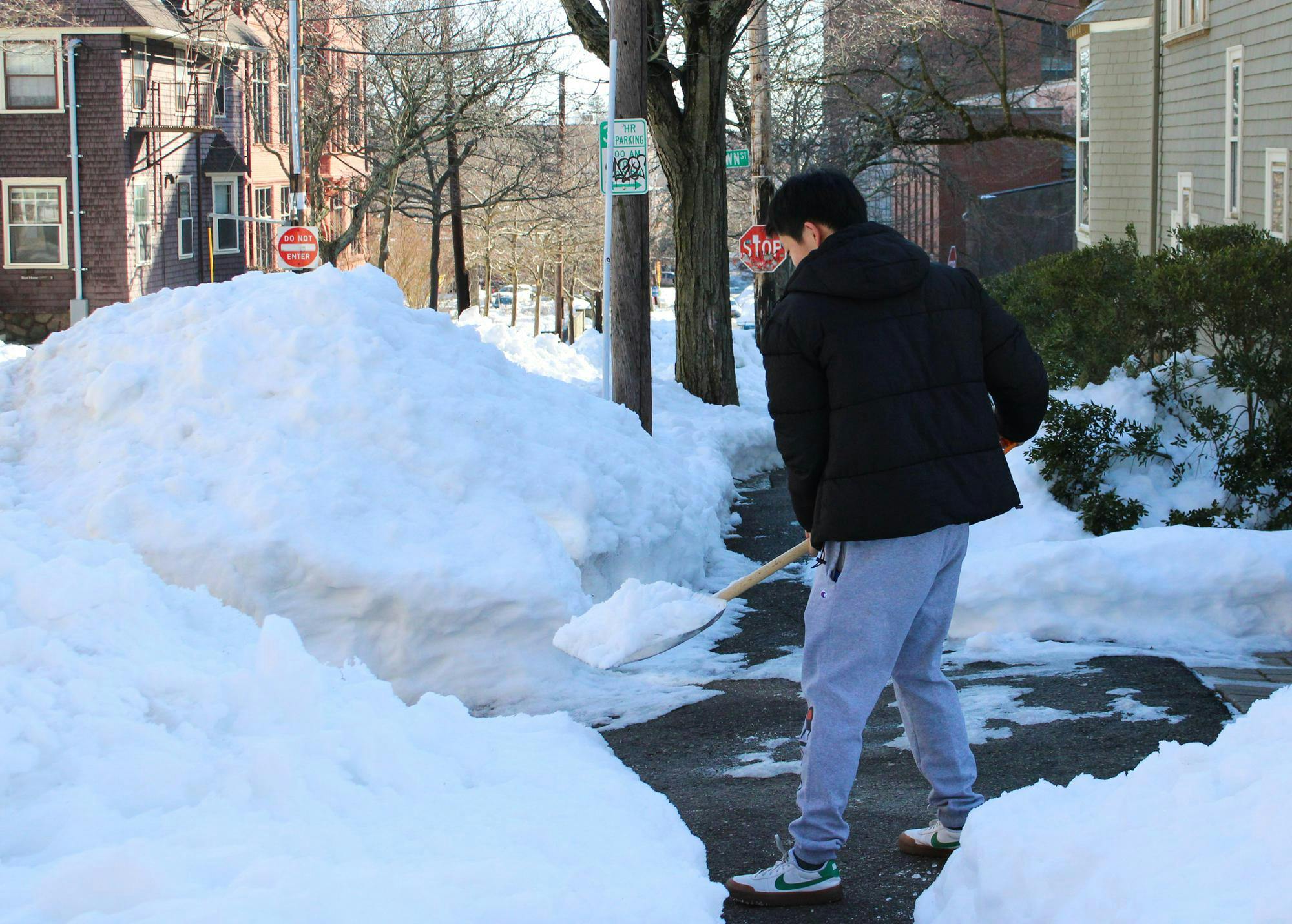 A person shoveling snow off a partially cleared sidewalk into a nearby pile of snow.