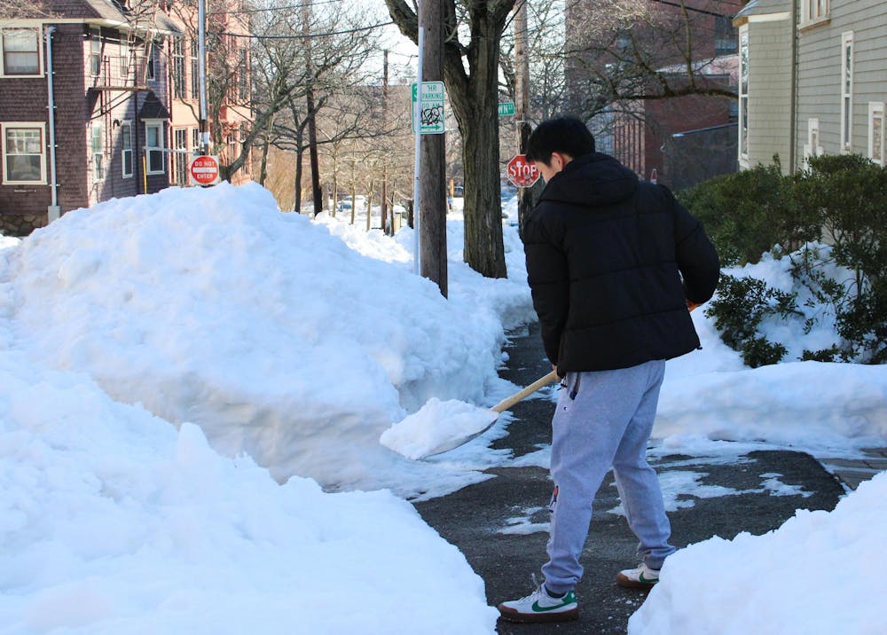 A person shoveling snow off a partially cleared sidewalk into a nearby pile of snow.
