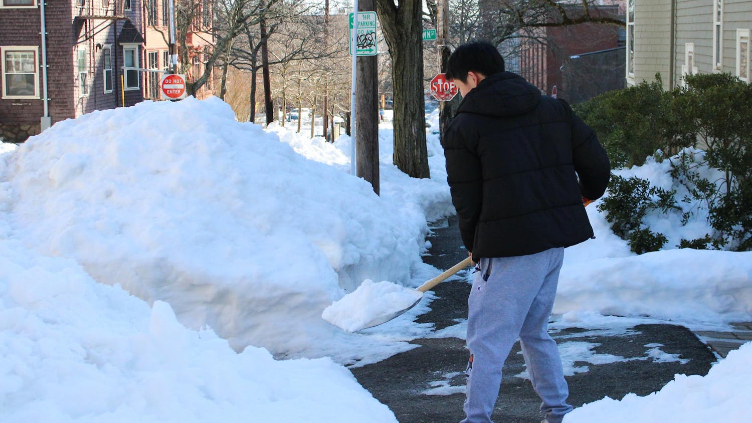 A person shoveling snow off a partially cleared sidewalk into a nearby pile of snow.
