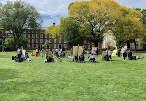 A circle of students paint on large canvases in front of the elm tree on the Main Green. The leaves are green, tinged with yellow. 