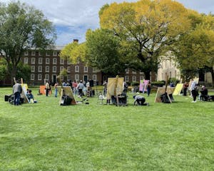 A circle of students paint on large canvases in front of the elm tree on the Main Green. The leaves are green, tinged with yellow.