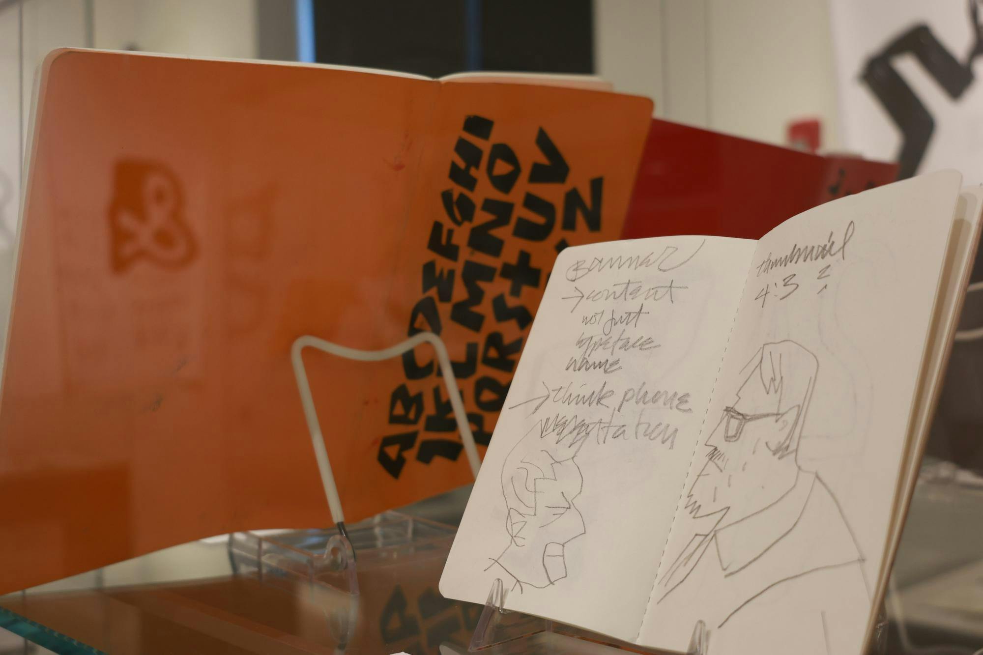 Photo of a sketchbook viewed in a clear display case. The sketchbook is open to a page featuring a drawing of someone viewed in side profile and wearing glasses, with various notes and another face sketch on the opposite page.