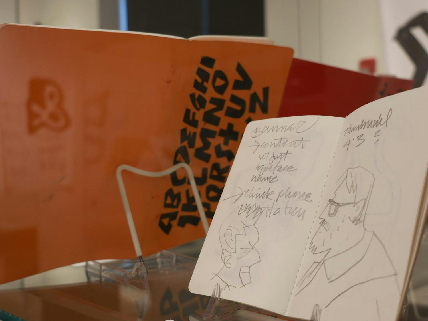Photo of a sketchbook viewed in a clear display case. The sketchbook is open to a page featuring a drawing of someone viewed in side profile and wearing glasses, with various notes and another face sketch on the opposite page.