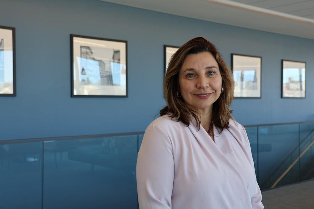 A picture of Sandra Smith in front of a blue wall with several small printed photographs decorating the wall. Sandra is wearing a pink top and smiling at the camera.