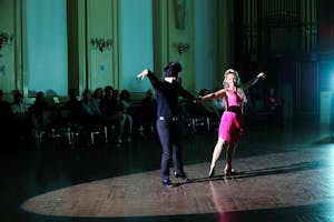 A woman in a pink dress and a man in all black dance in a large ballroom.