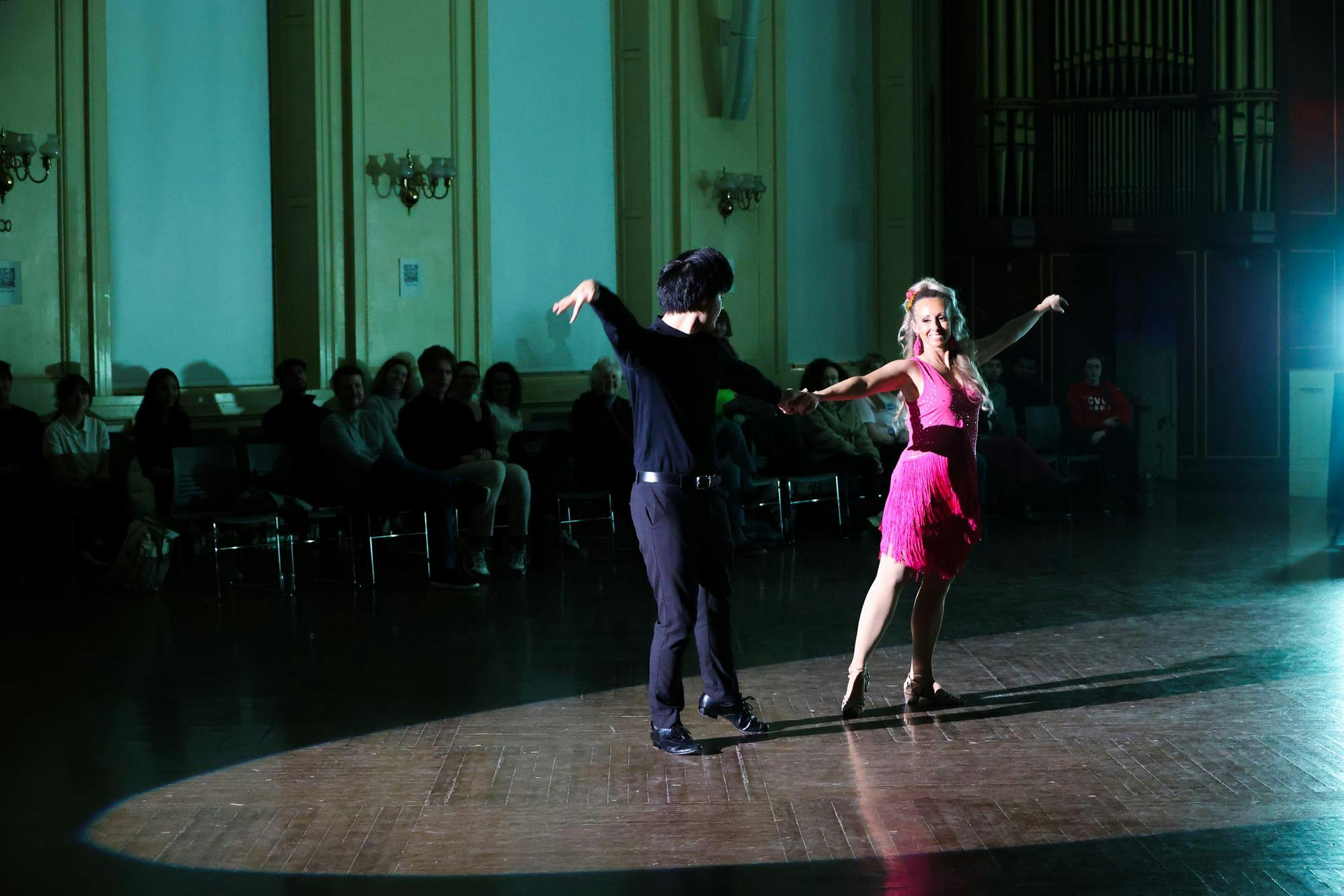 A woman in a pink dress and a man in all black dance in a large ballroom.