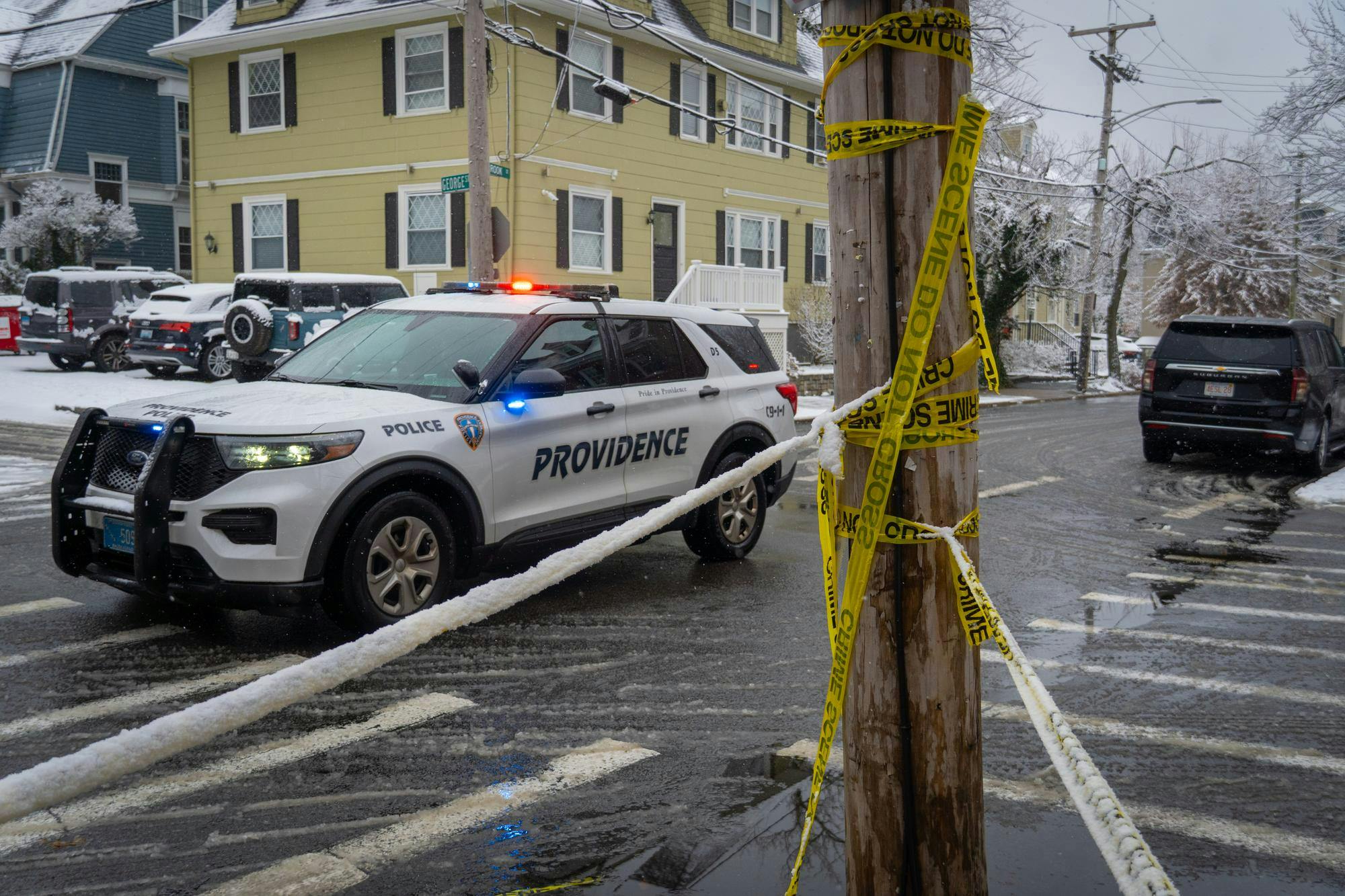 A photo of yellow caution tape wrapped around a post in front of a street intersection with a police car driving past. 