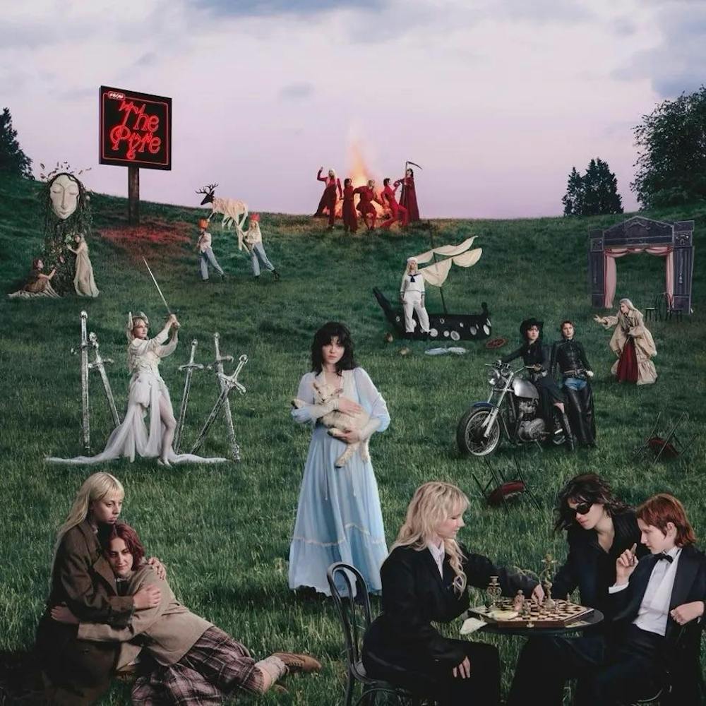 Album cover for ‘From the Pyre’ by The Last Dinner Party, which shows a series of figrures and scenes placed disparately around a grassy hill. The central figure, a woman in a blue dress, holds a lamb. At the top of the hill, several figures dressed in red move around a burning pyre.