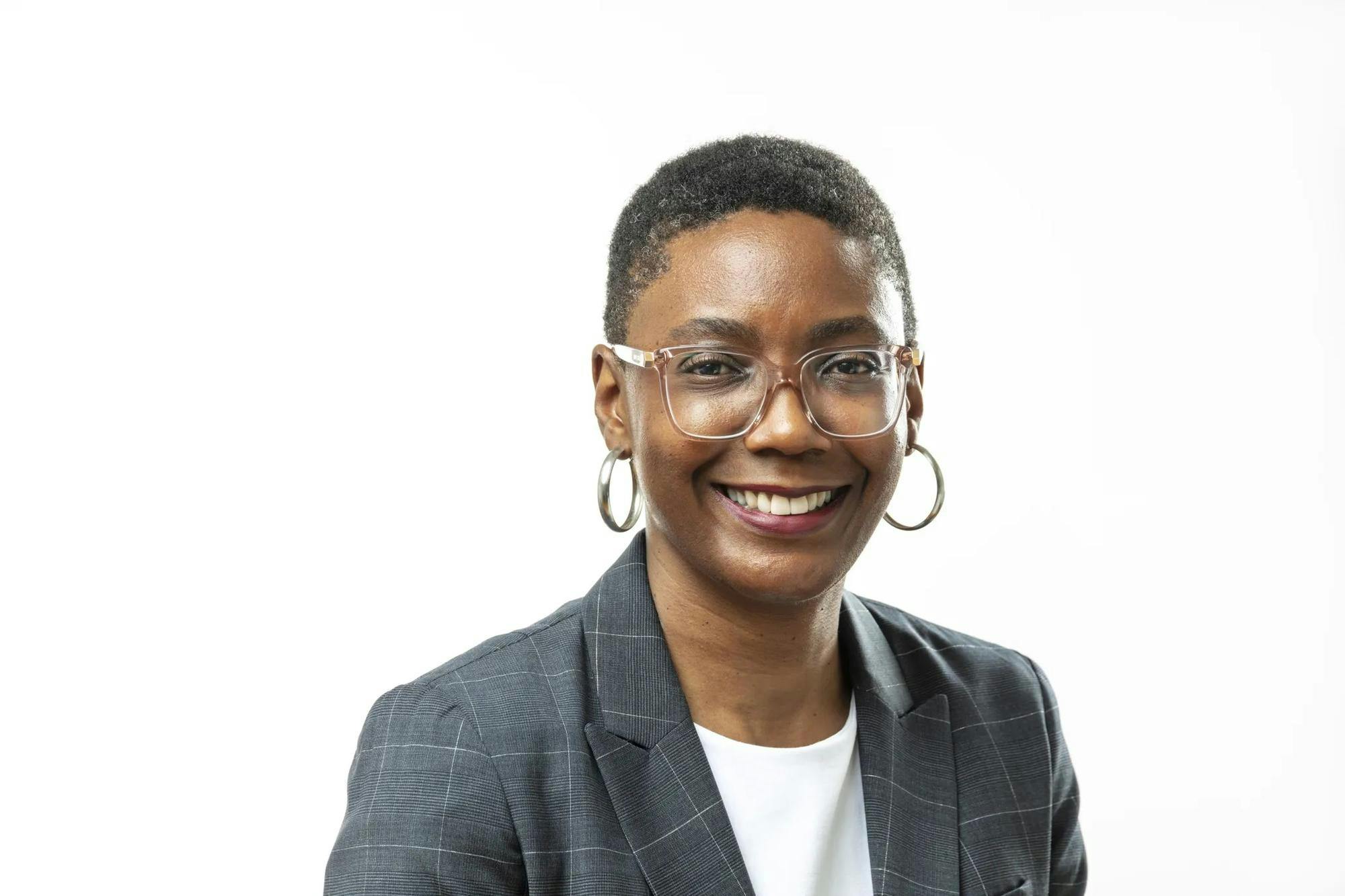 Photo of a woman in a grey checkered blazer and white shirt, wearing silver hoop earrings and a pair of clear glasses.