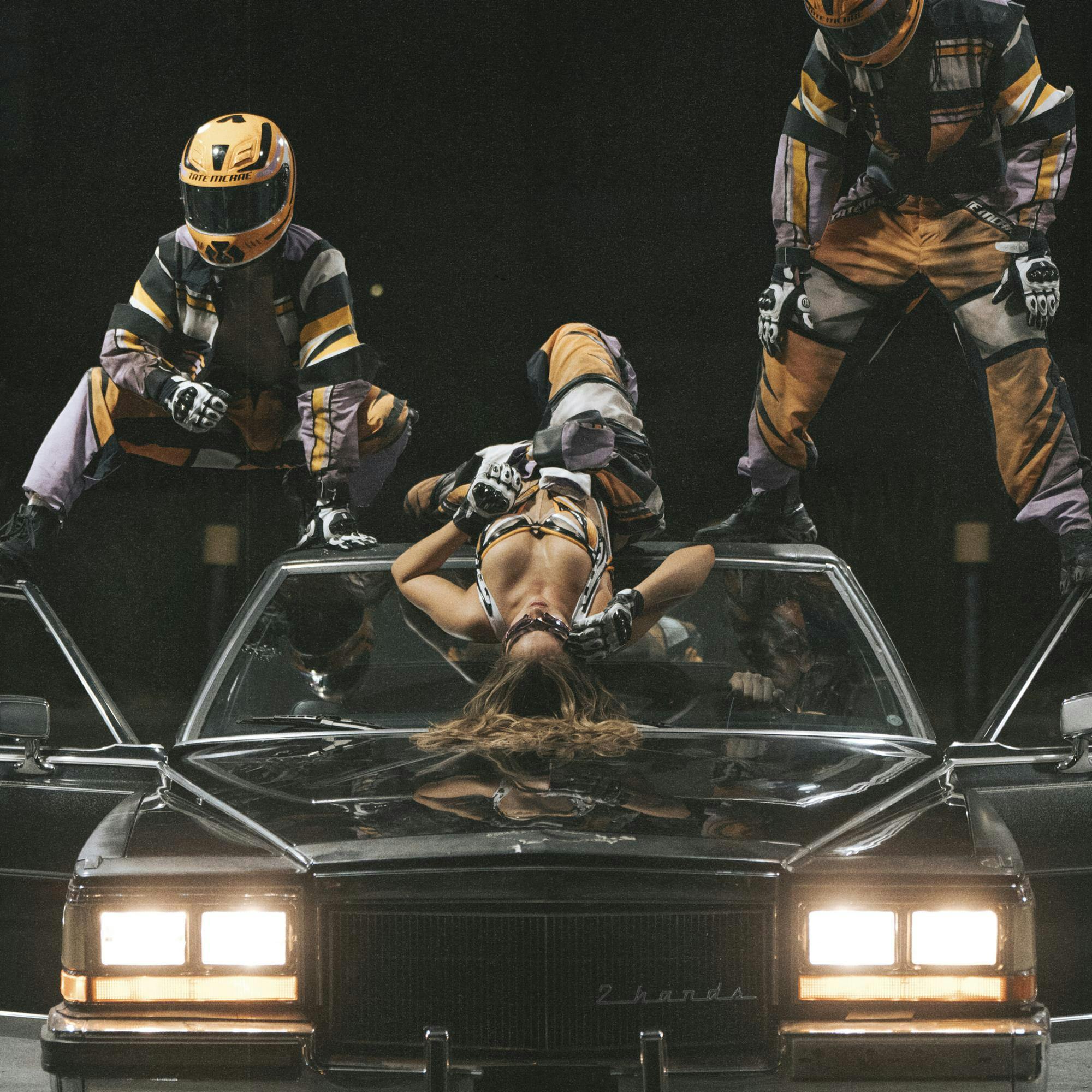 A woman lies upside down on the roof of a car with two people standing at either side.