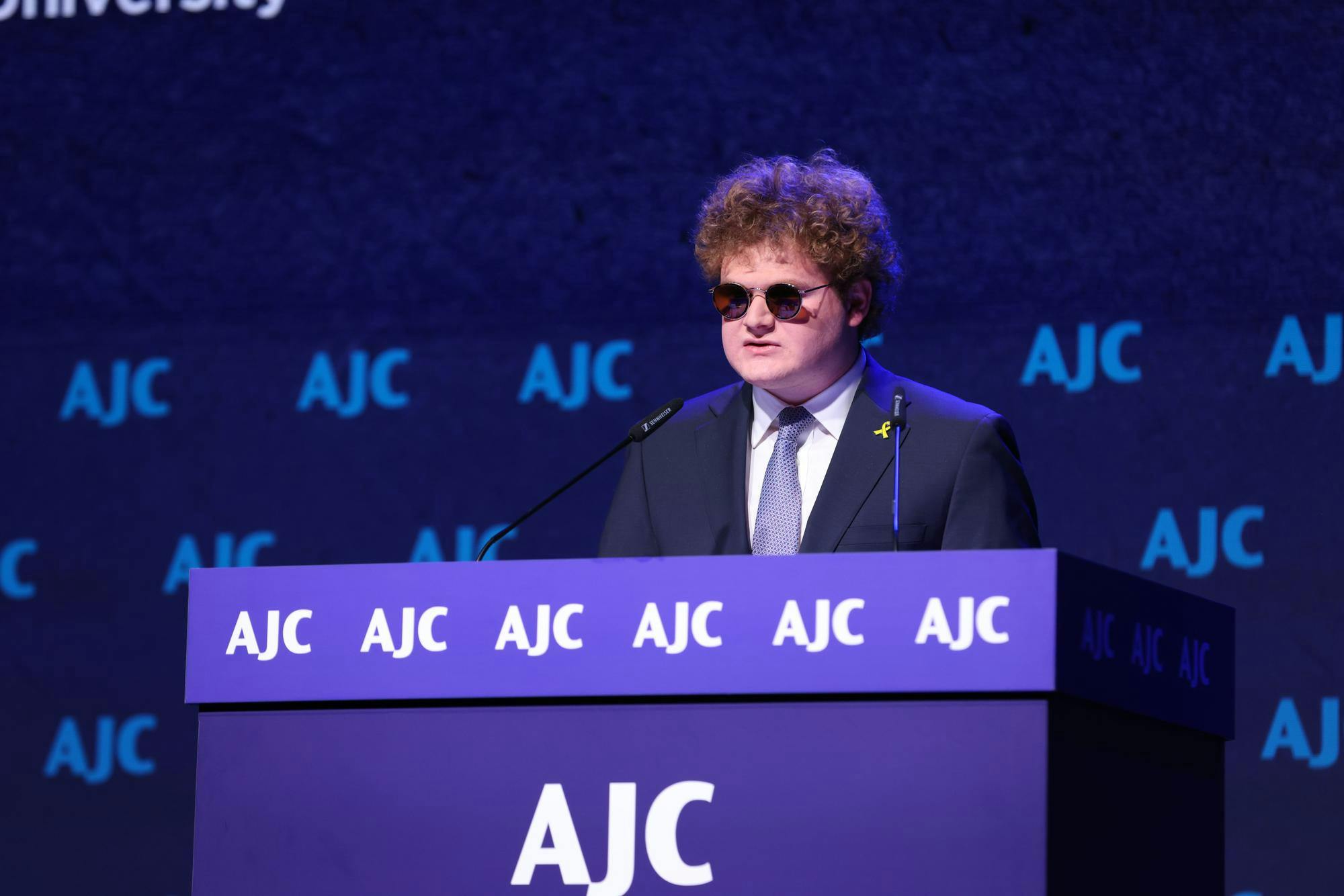 A photo of Daniel Solomon '26 delivering remarks at the AJC Global Forum in NYC. He is speaking at the podium, wearing a suit with a yellow ribbon.