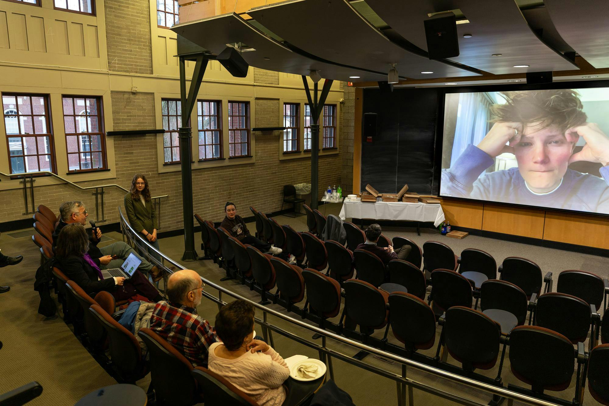 A photo of a lecture hall with a large zoom screen displaying Charlie Covell's face. 