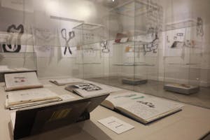 Photo of the “Everywhere I Go,” exhibit at the Providence Public Library on Tuesday, featuring sketchbooks open and on display in clear display cases with various larger drawings displayed on the walls.