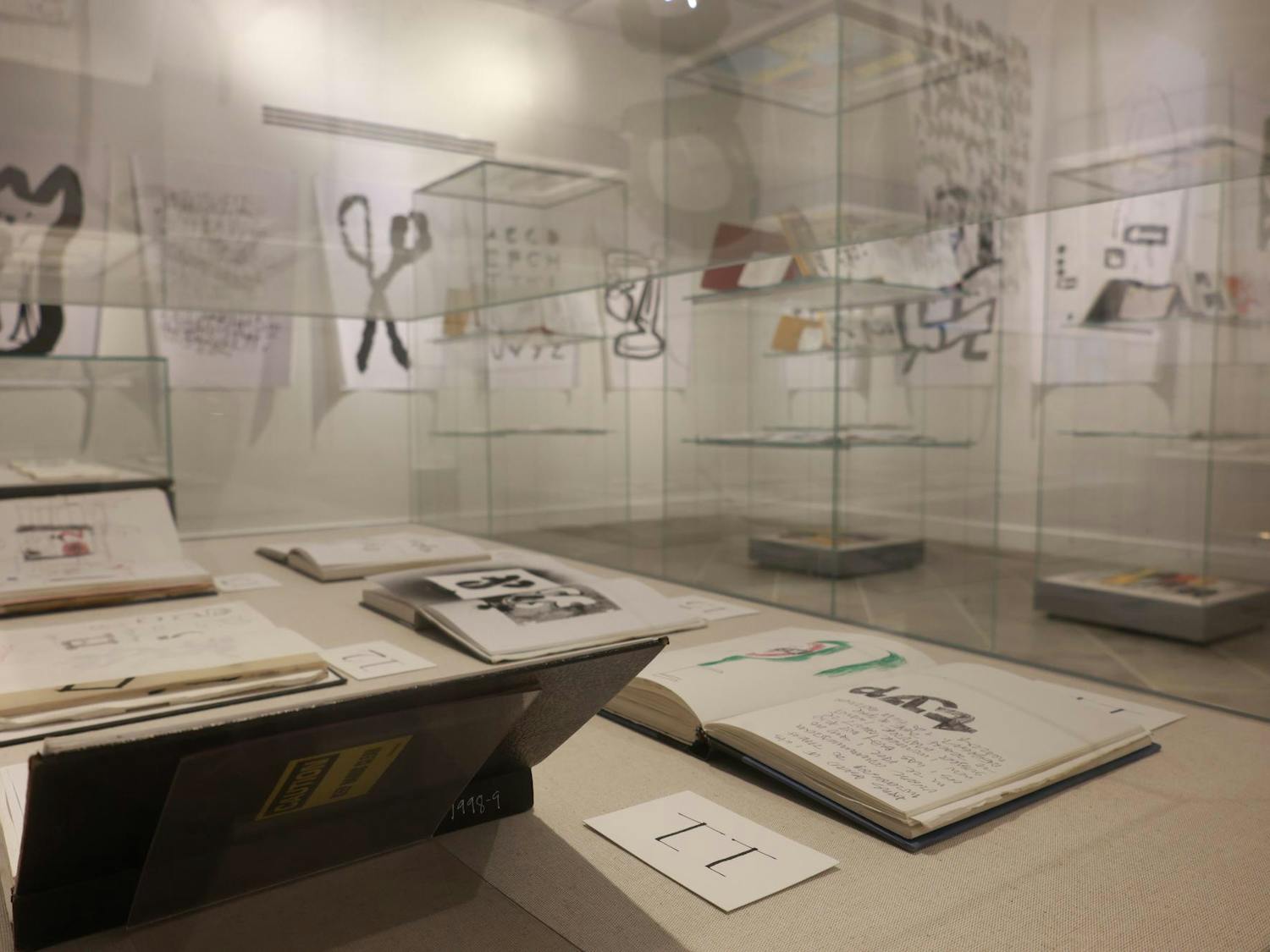 Photo of the “Everywhere I Go,” exhibit at the Providence Public Library on Tuesday, featuring sketchbooks open and on display in clear display cases with various larger drawings displayed on the walls.