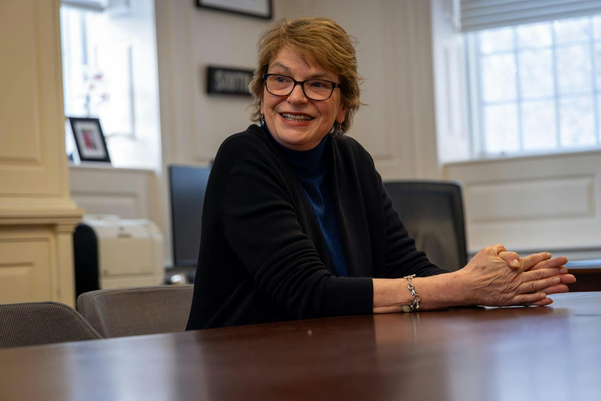 Paxson looks just past the camera while leaning over a table in a blue shirt and black sweater.