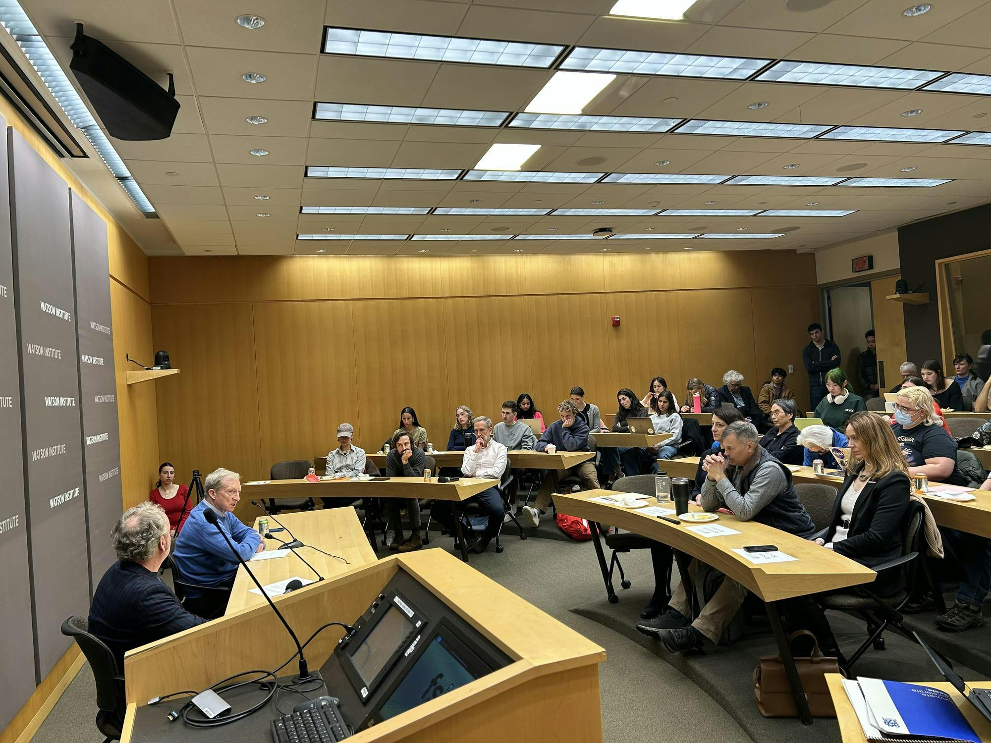 Approximately 30 people sit watching a man in a blue sweater speaking. 