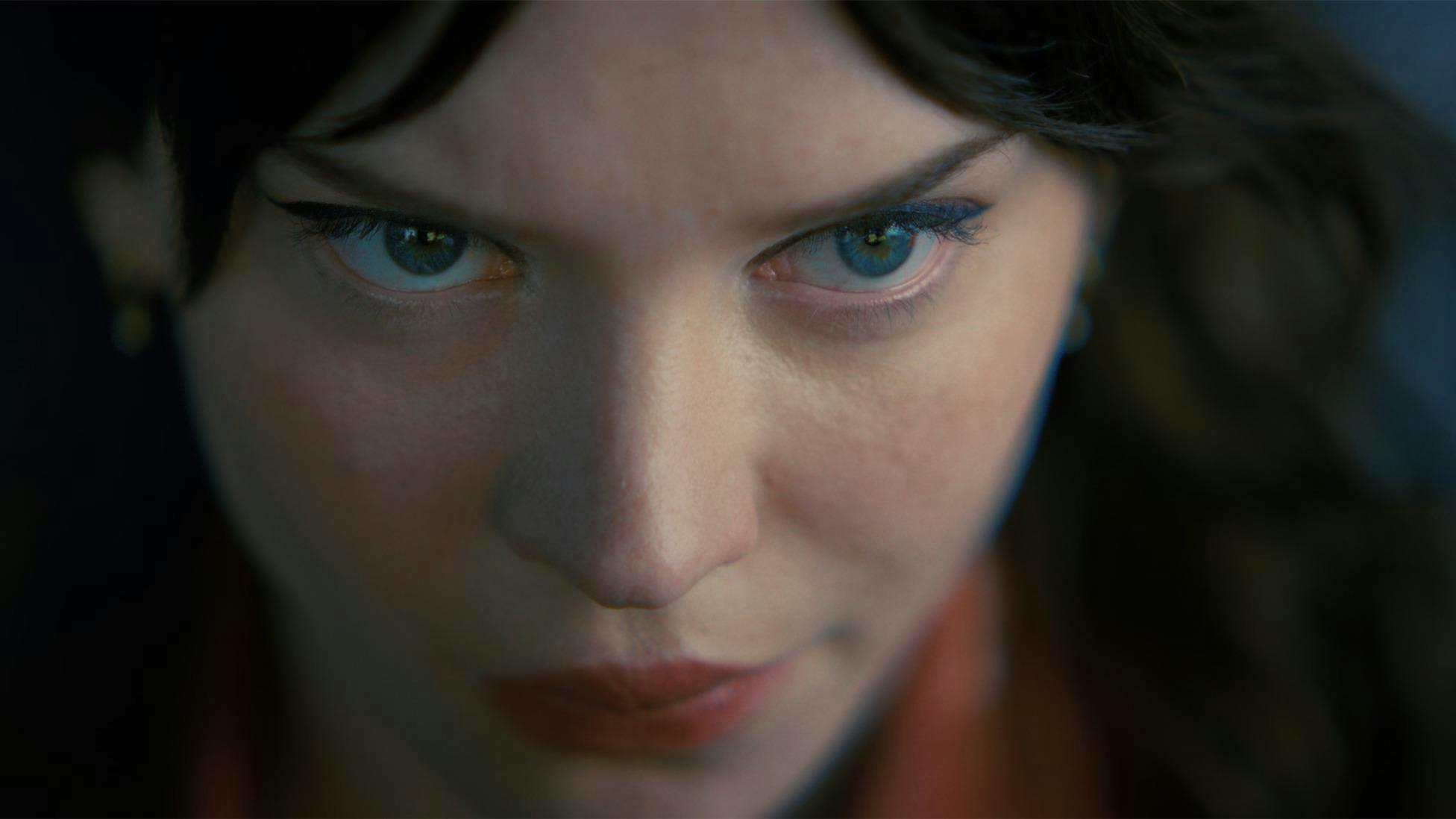 Close up shot of Sophie Thatcher playing Iris in "Companion". Her eyes glitter a brilliant blue beneath teal eyeshadow. Red lipstick colors her smirk.