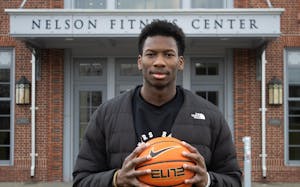 A photo of a basketball player standing in front of the Nelson Fitness Center wearing a black North Face jacket holding a basketball in front of him.