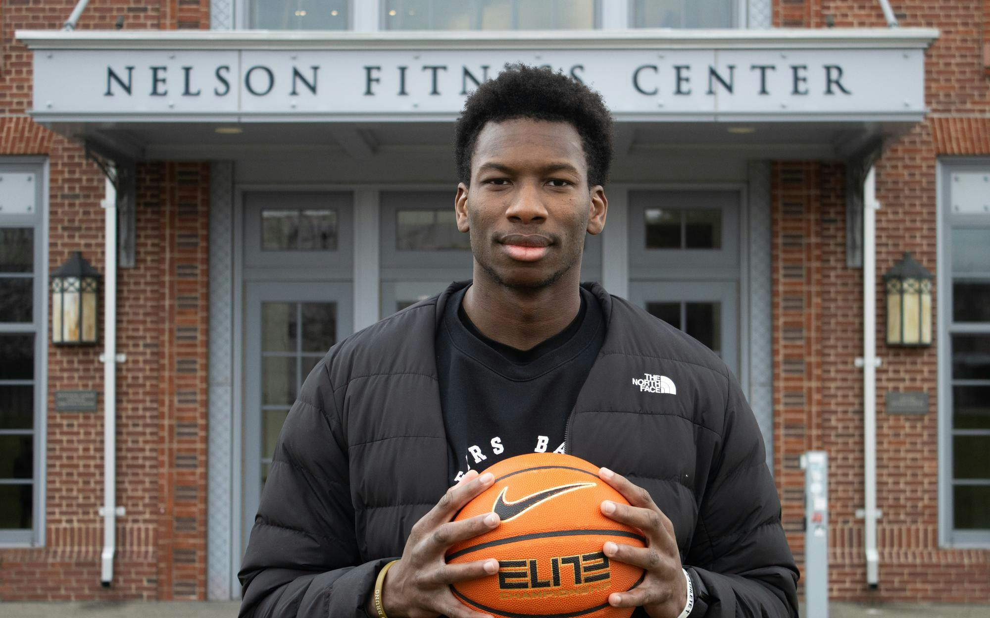 A photo of a basketball player standing in front of the Nelson Fitness Center wearing a black North Face jacket holding a basketball in front of him.