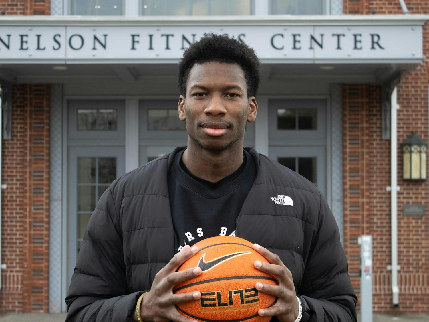 A photo of a basketball player standing in front of the Nelson Fitness Center wearing a black North Face jacket holding a basketball in front of him.
