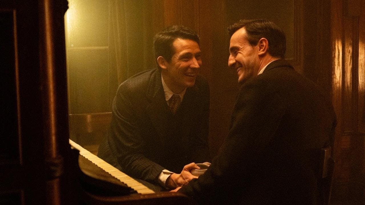 In a brown hazy photo, two men in suits laughing with each other and holding hands while sitting on a piano together.