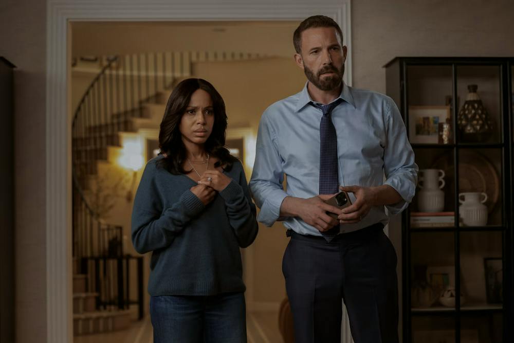 Photo of a woman wearing a grey sweater and jeans, standing looking worried while clutching her necklace. A man holding a phone is on the right wearing a dress shirt, tie and dress pants.