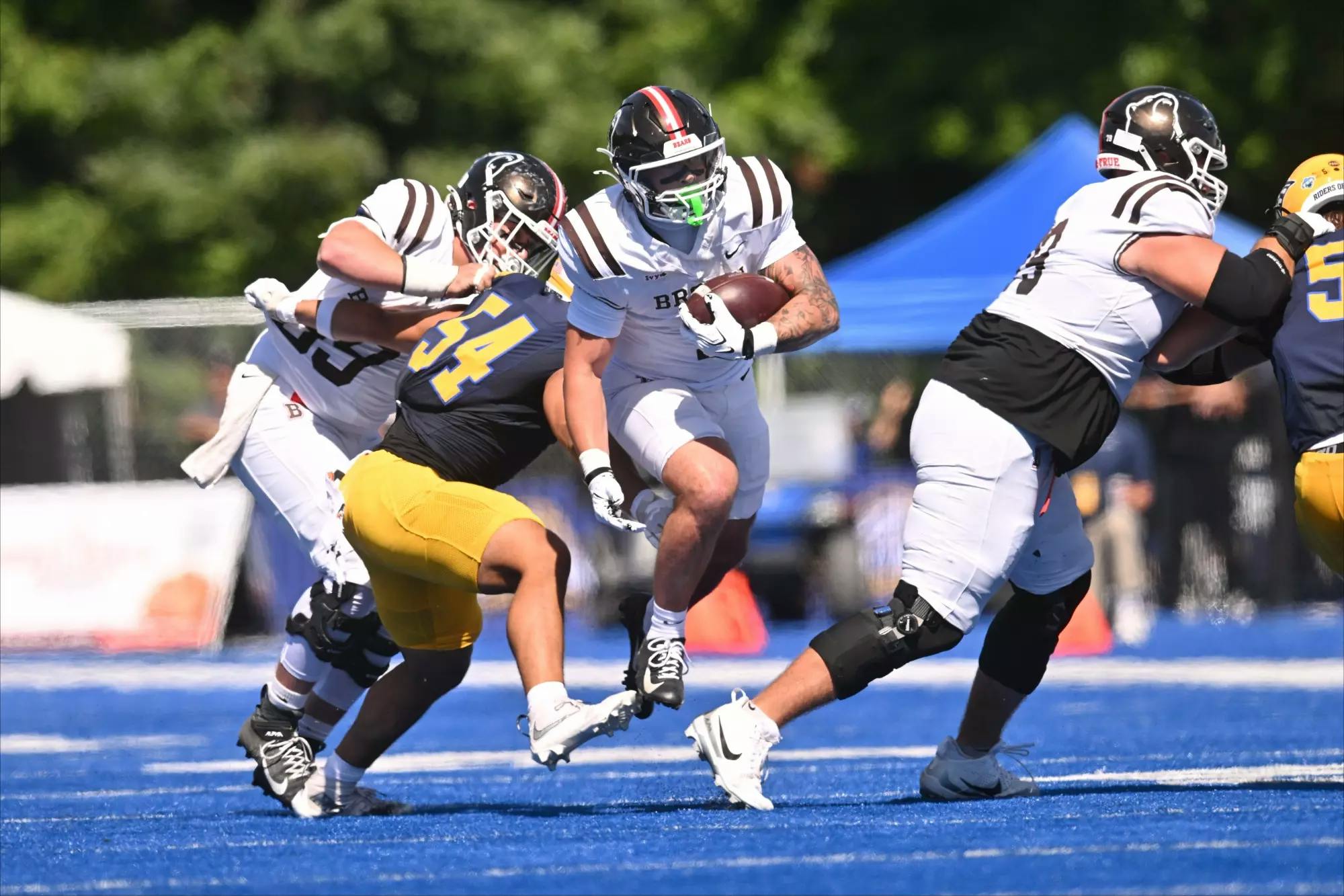 Photo of four football players, one player for New Haven tackles two Brown University players. The Brown University player on the right is cradling the ball. One more Brown University player tackles a New Haven player on the right.