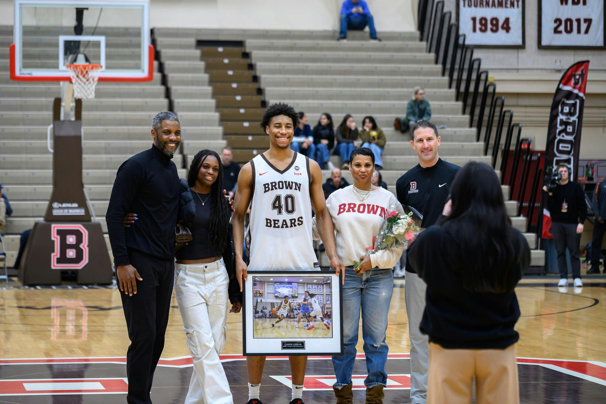 Lewis stands on the basketball court with his mother, father, and sister with him. He is holding a plaque with a picture of him during a game.