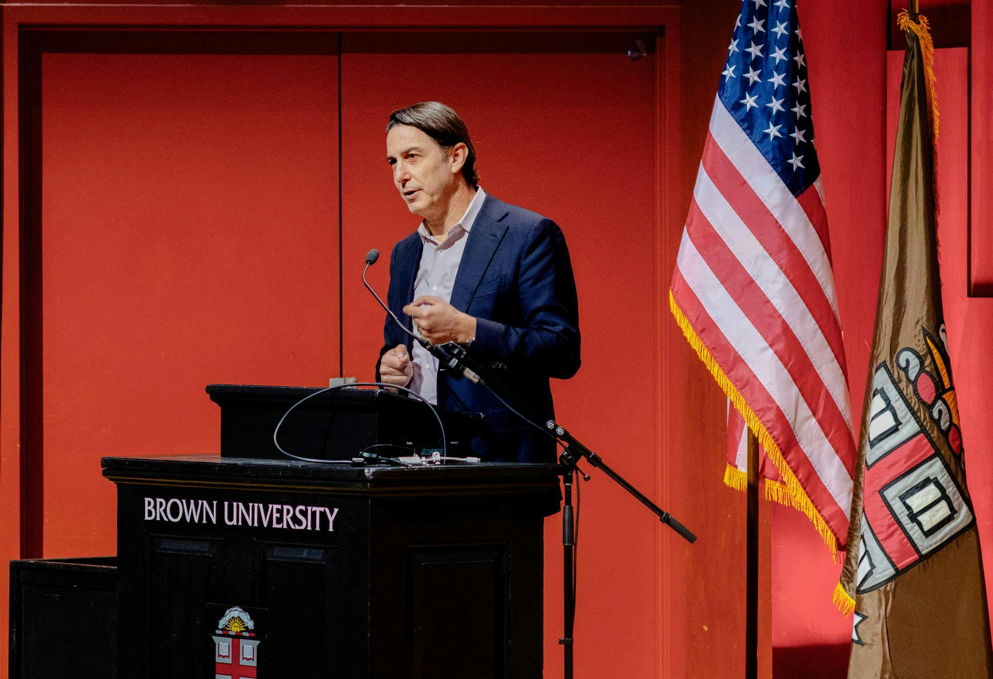 Amos Hochstein wears a navy blue suit and stands behind a black Brown University podium in front of a red wall. 