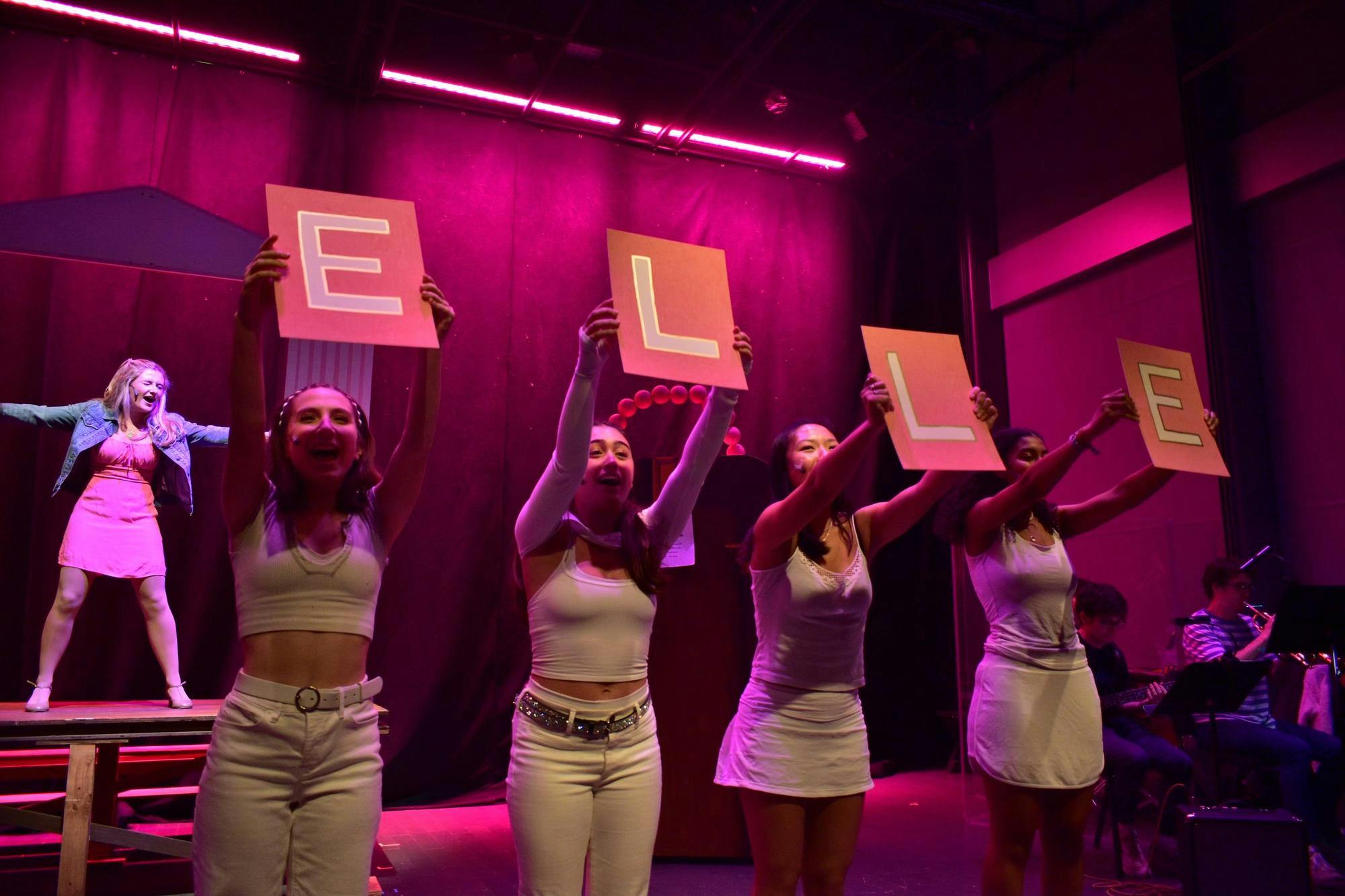 Four actors dressed in white hold up signs that spell out Elle, as Elle Woods sings in the background. 