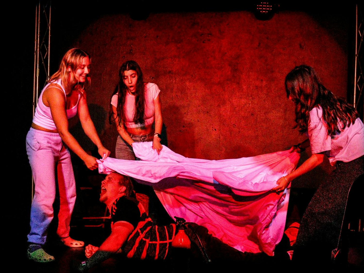 Three actors appear to be smothering a fourth with a sheet on a darkly lit stage.
